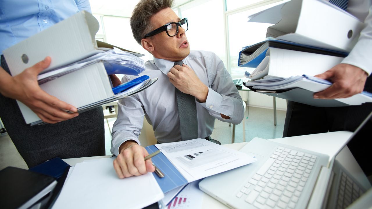 Two people are giving a worker other documents to work on while he is overloaded with existing papers on the desk.